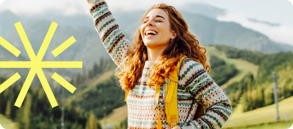 Happy woman with curly hair wearing a patterned sweater and yellow backpack outdoors in a mountainous landscape.