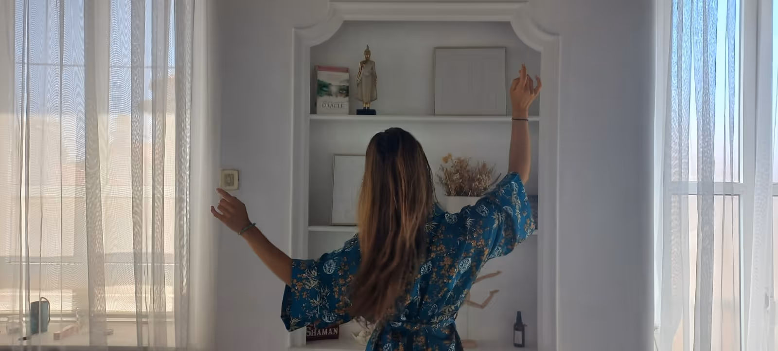 Mulher de cabelos longos e vestido azul com padrão floral, de costas, com os braços levantados num quarto iluminado pela luz natural através de cortinas brancas.