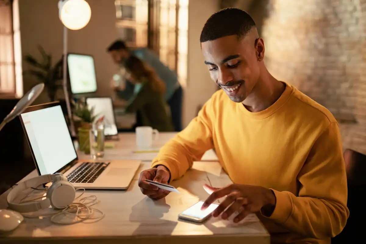 Homem sorridente usando smartphone e cartão de crédito em uma mesa de trabalho moderna, com laptop e colegas ao fundo.