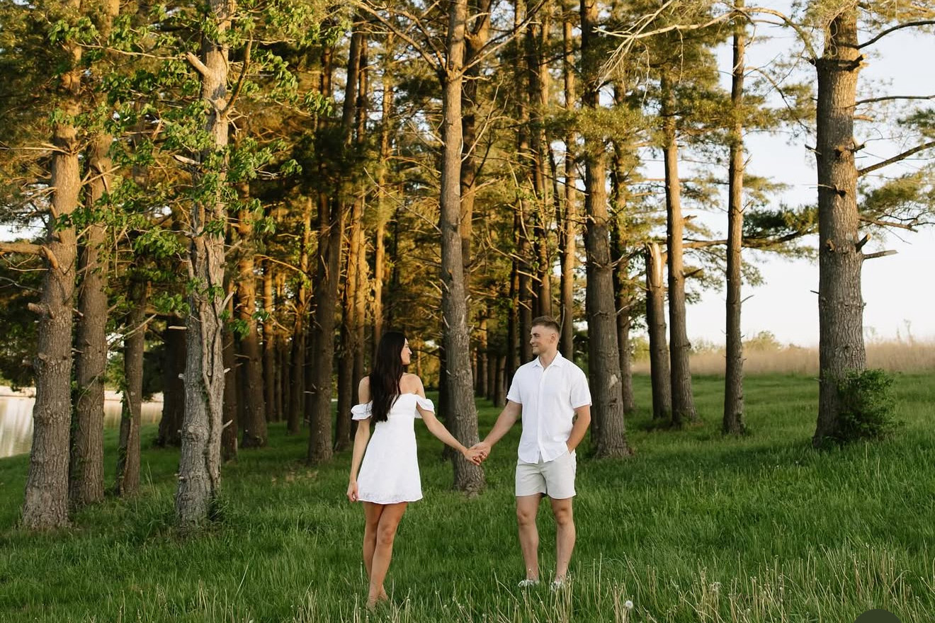 A couple doing an enegagement photo shoot with a treeline behind them at the Gathering Grounds