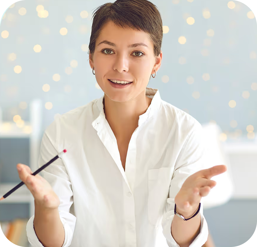 Smiling woman in white shirt gesturing with hands while holding a pencil in a softly lit room.