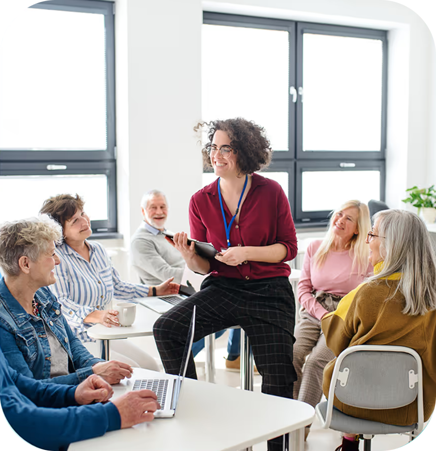 A woman with curly hair and glasses leading a discussion with a group of older adults in a bright room.