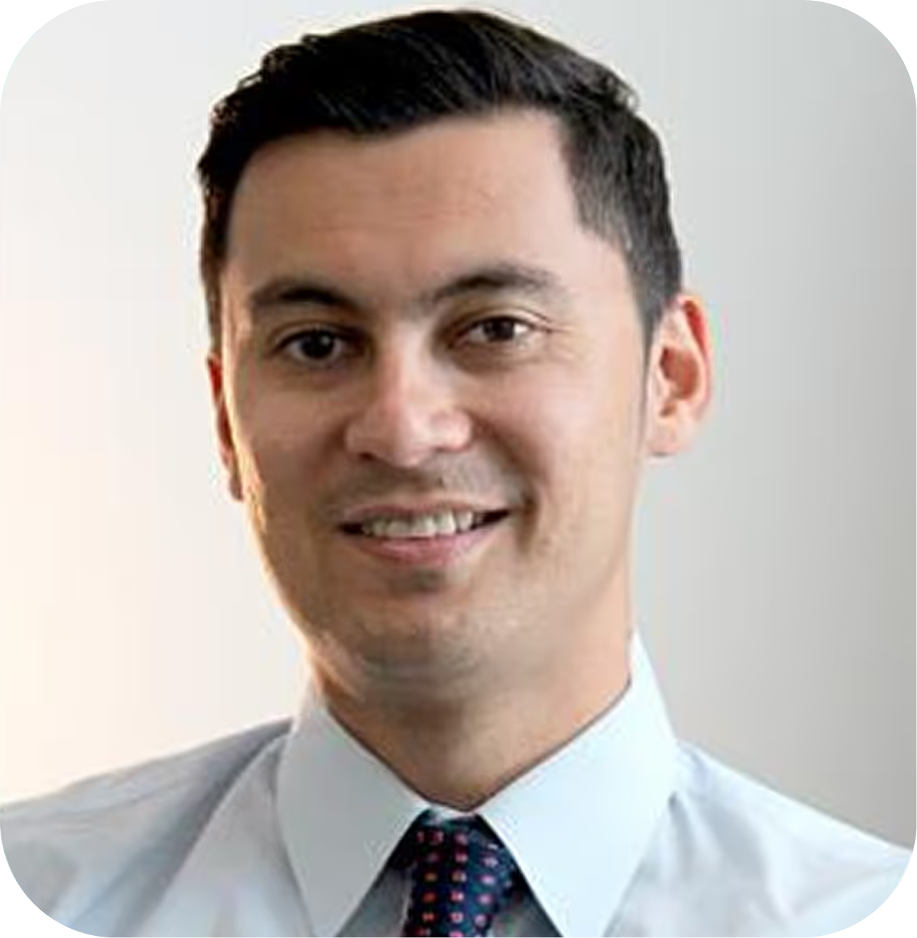 Smiling man with dark hair wearing a light blue shirt and polka dot tie against a plain background.