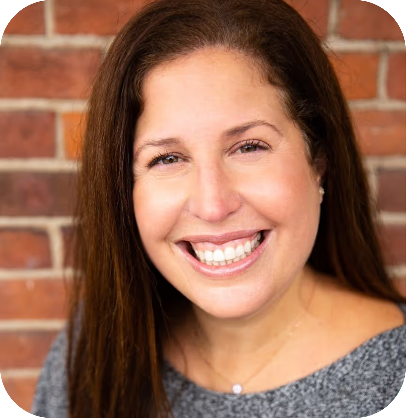 Smiling woman with long brown hair, wearing a gray top and a delicate necklace, in front of a brick wall.