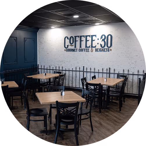 Modern coffee shop interior with wooden tables, black chairs, and a white brick wall featuring the sign 'COFFEE 30 GOURMET COFFEE & BEIGNETS.'