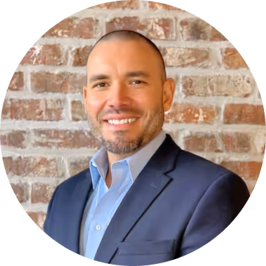 Smiling man with short beard and closely shaved hair wearing a navy blue suit jacket and light blue shirt in front of a brick wall.
