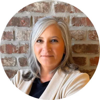 Smiling middle-aged woman with gray hair wearing a beige cardigan and black top, standing in front of a brick wall.