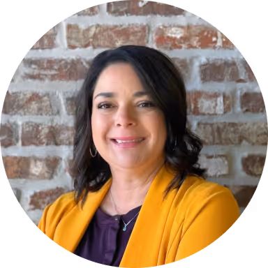 Smiling woman with dark hair wearing a mustard yellow cardigan and purple top in front of a brick wall.