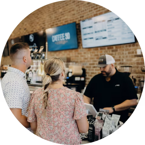 Two customers standing at a coffee shop counter ordering from a barista wearing a cap.