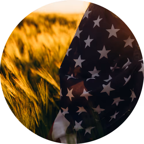 Person draped in an American flag standing in a golden wheat field at sunset.