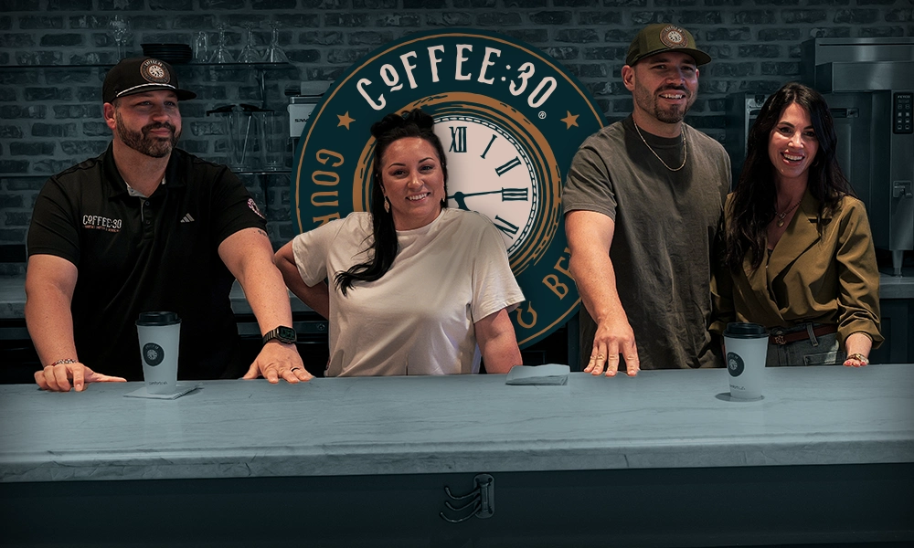 Four smiling people standing behind a marble counter with coffee cups, with a Coffee:30 logo on the brick wall behind them.