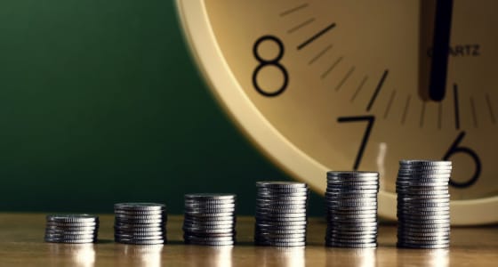 Several stacks of coins increasing in height from left to right with a clock in the background