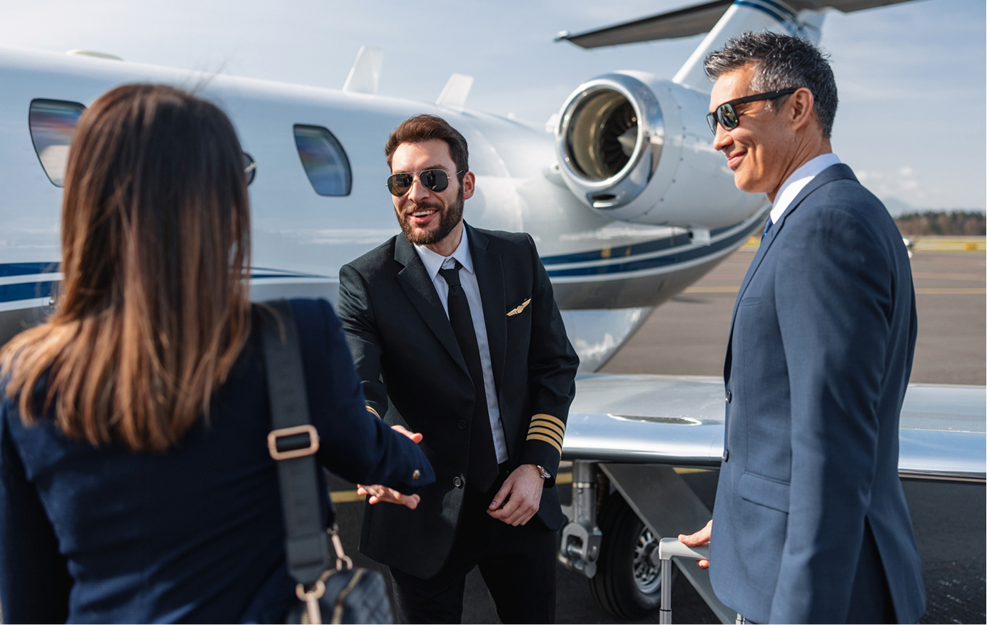Un pilote en uniforme serre la main d'une passagère près d'un avion d'affaires privé tandis qu'un homme en costume tient une valise à roulettes.