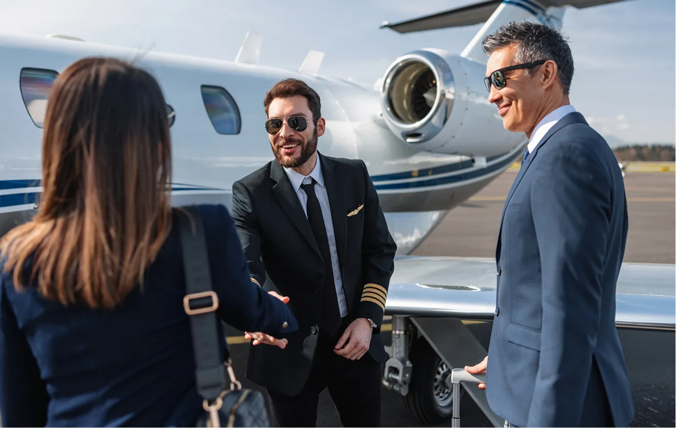 Un pilote en uniforme serre la main d'une passagère près d'un avion d'affaires privé tandis qu'un homme en costume tient une valise à roulettes.