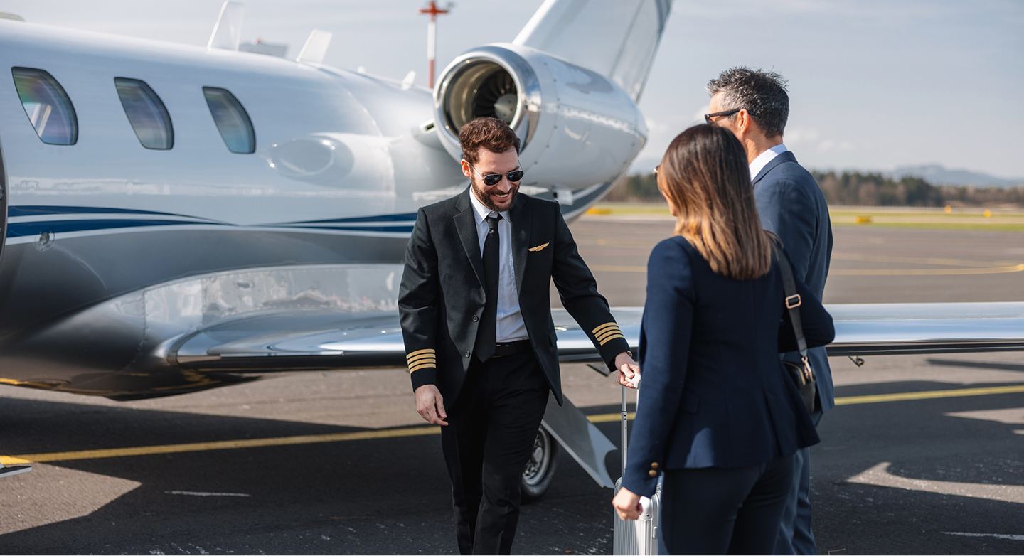 Un pilote souriant en uniforme noir accueille deux passagers vêtus de costumes devant un petit jet privé à l'aéroport.