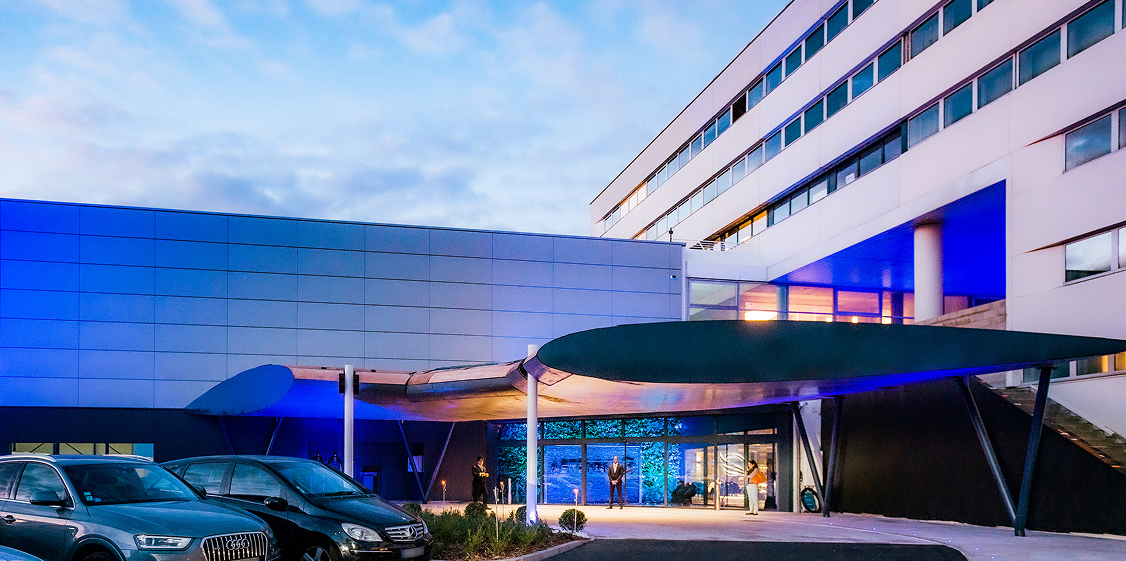 Entrée moderne d'un bâtiment avec auvent incurvé, voitures stationnées et lumière bleue illuminant la façade au crépuscule.