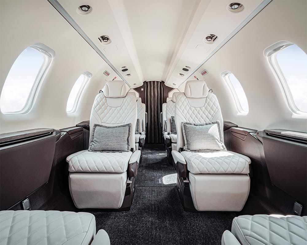 Intérieur luxueux d'un jet privé avec des sièges en cuir blanc matelassé et des coussins gris sous les fenêtres.