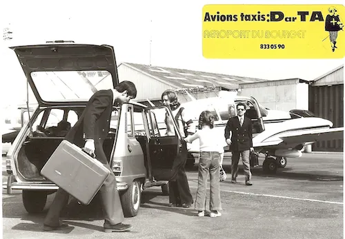 Un homme portant une valise ouvre le coffre d'une voiture, une femme tient la porte arrière ouverte pour une autre femme et un enfant devant un petit avion sur un tarmac.