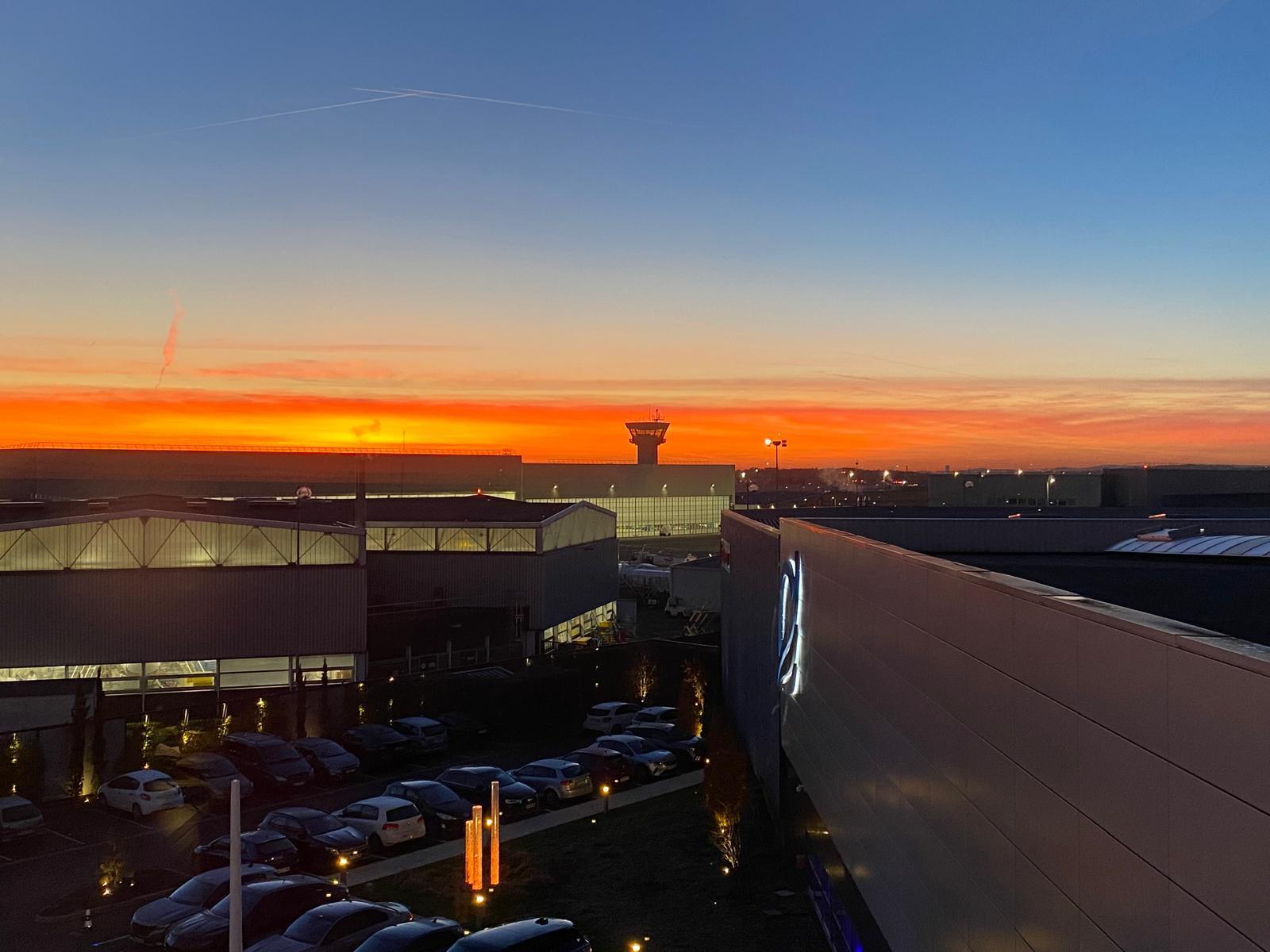 Coucher de soleil orangé au-dessus d'un parking rempli de voitures et de bâtiments industriels avec une tour de contrôle en arrière-plan.