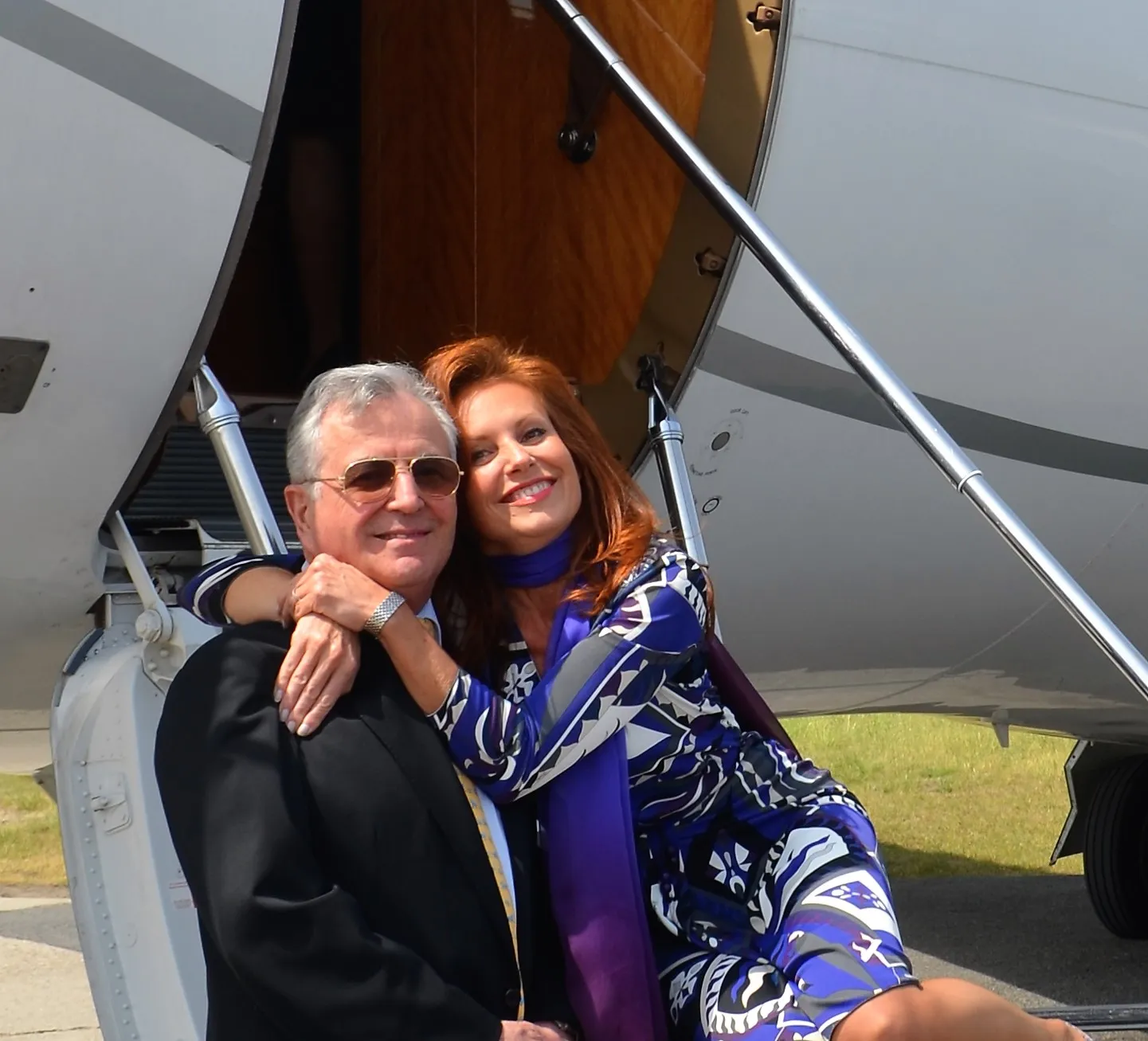 Un homme en costume noir avec des lunettes de soleil aux côtés d'une femme souriante portant une robe bleu et blanche, devant l'entrée d'un avion privé.