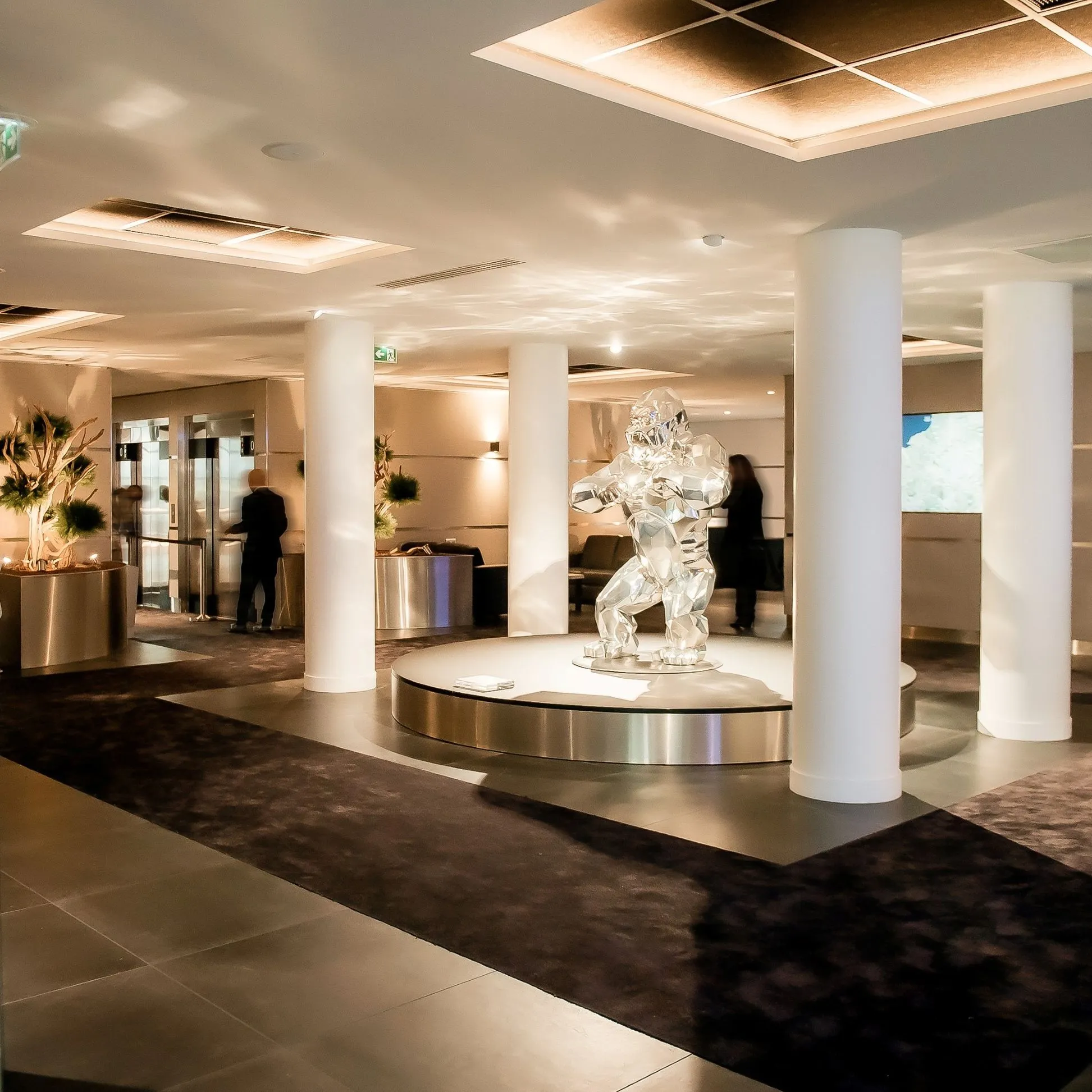 Hall d'entrée moderne avec une sculpture d'un gorille argenté sur un podium circulaire, entouré de colonnes blanches et d'un sol carrelé.
