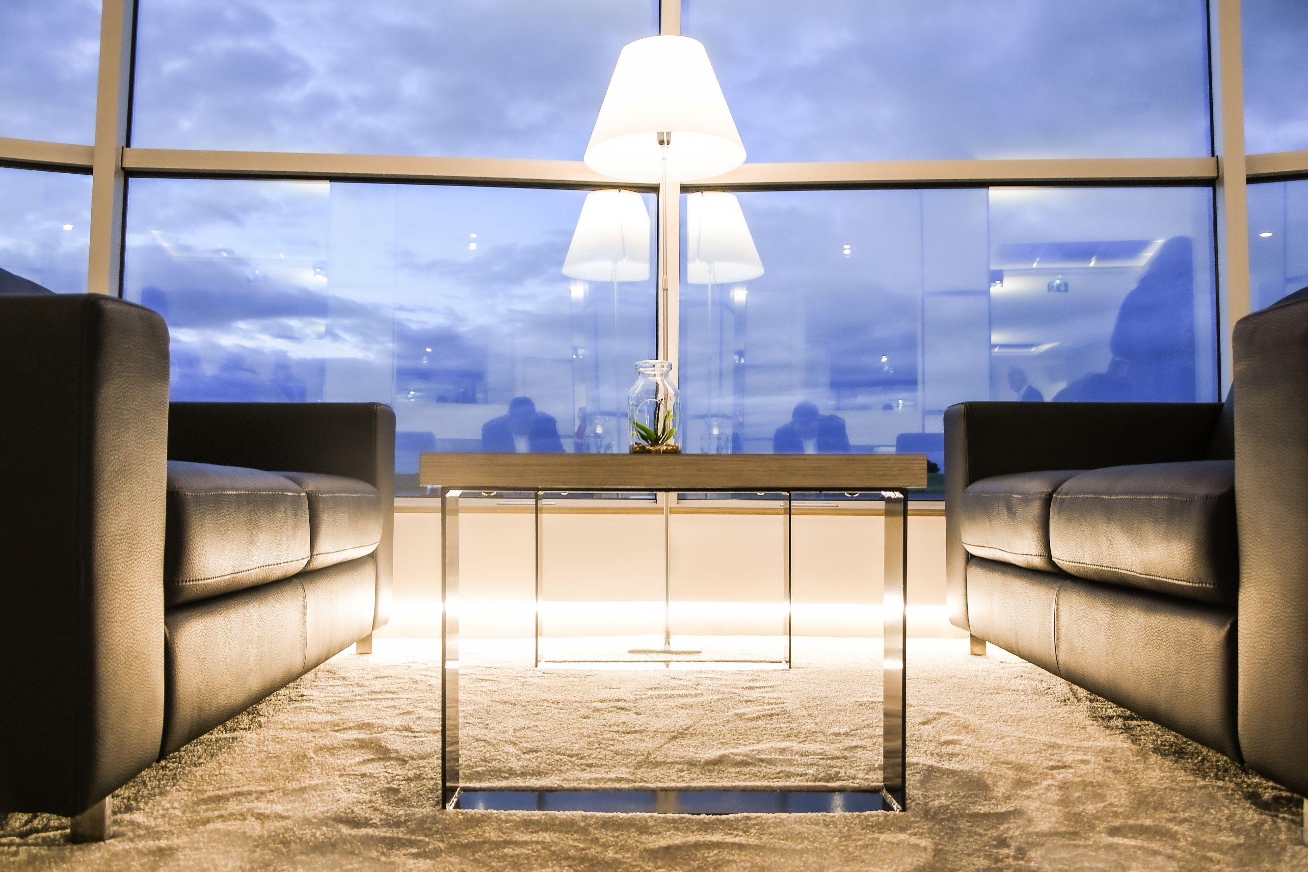 Deux canapés en cuir noirs face à face avec une table basse en métal et bois au centre, devant une grande fenêtre au crépuscule.