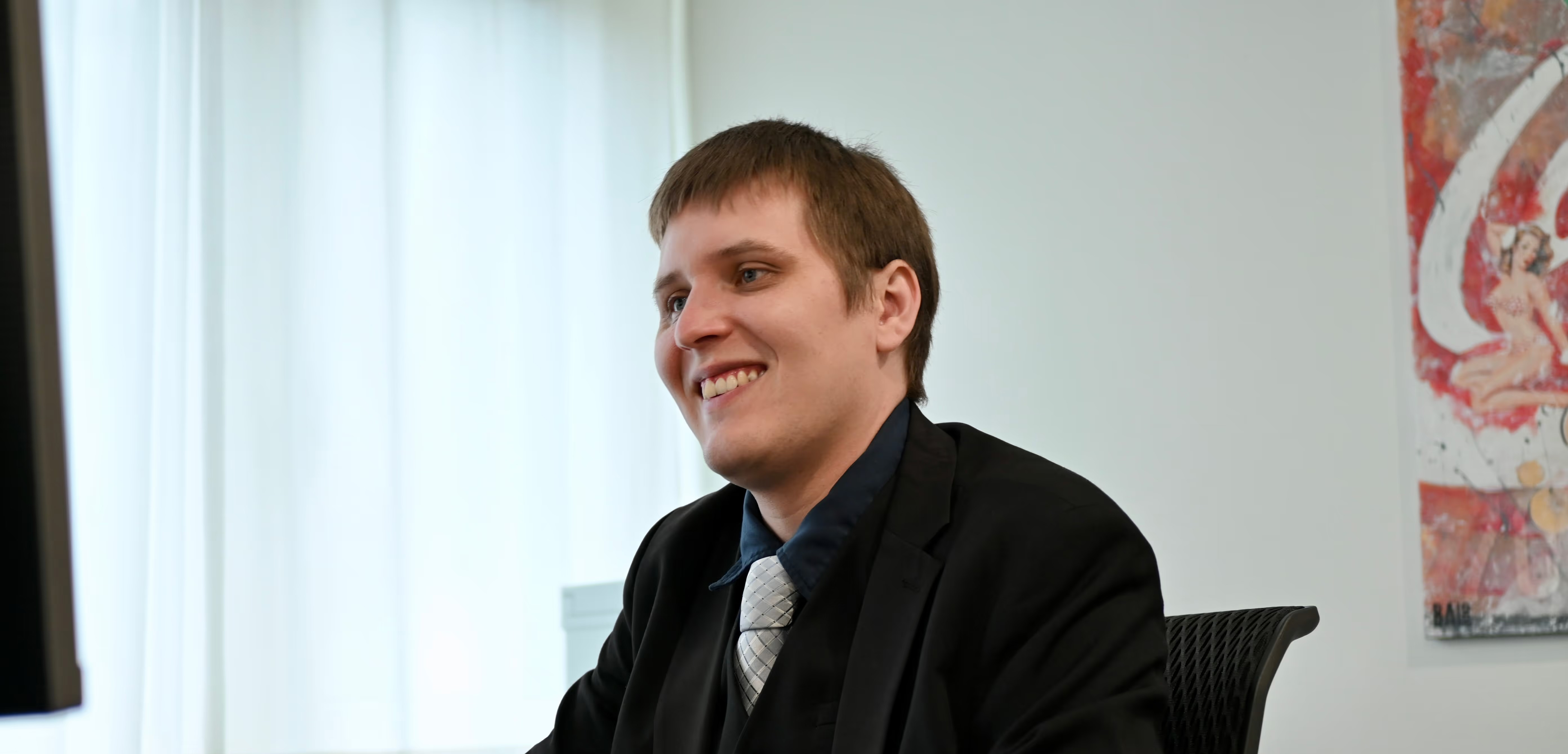 Ein Herr sitzt an seinem Arbeitsplatz und grinst in den Bildschirm.