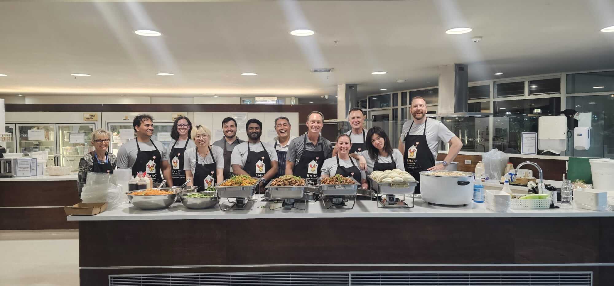 Group of volunteers in aprons standing behind a kitchen counter with trays of cooked food and utensils.