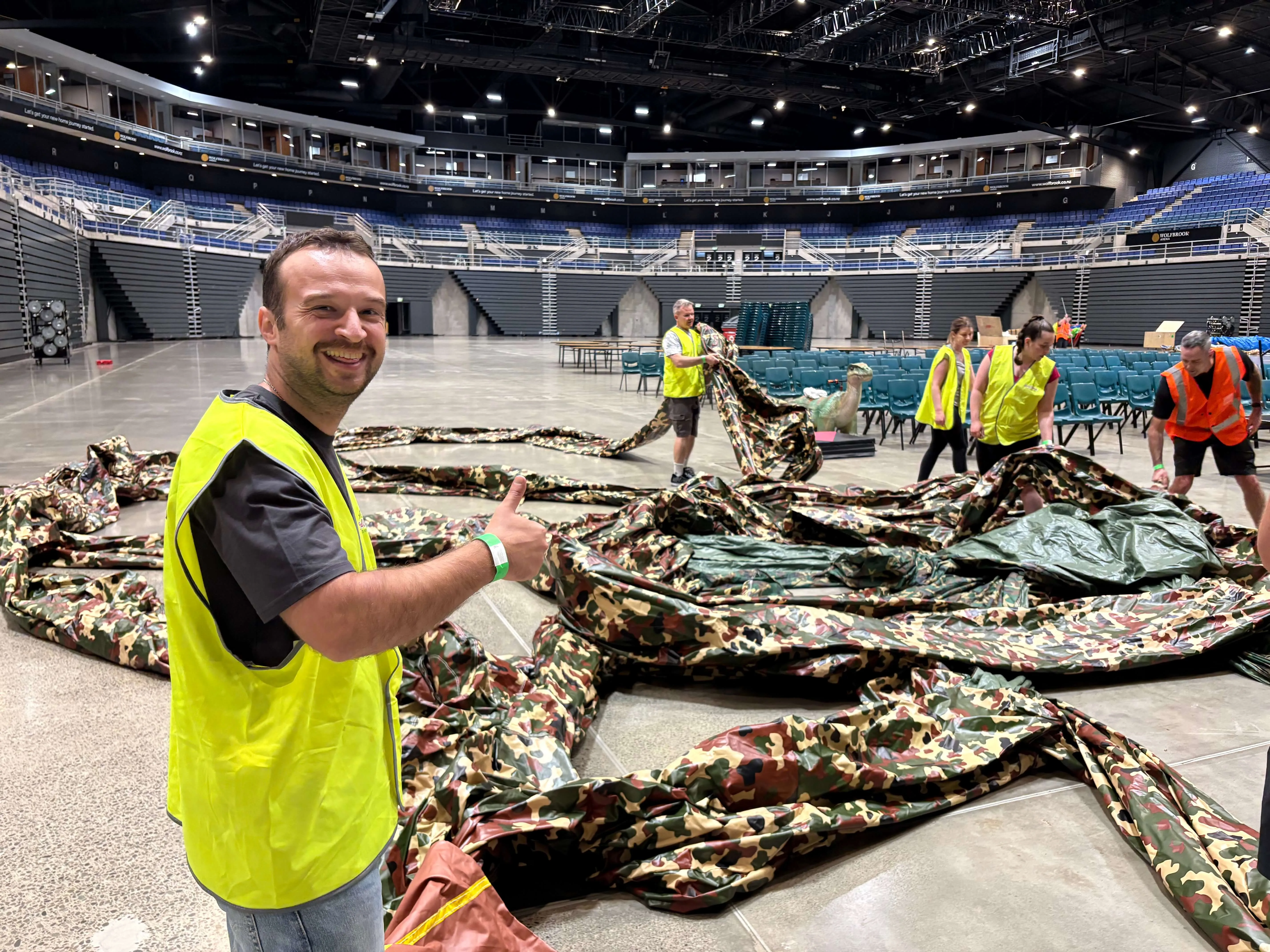 A smiling man in a yellow safety vest gives a thumbs-up inside a large indoor arena where people in safety vests arrange camouflage-patterned material on the floor.