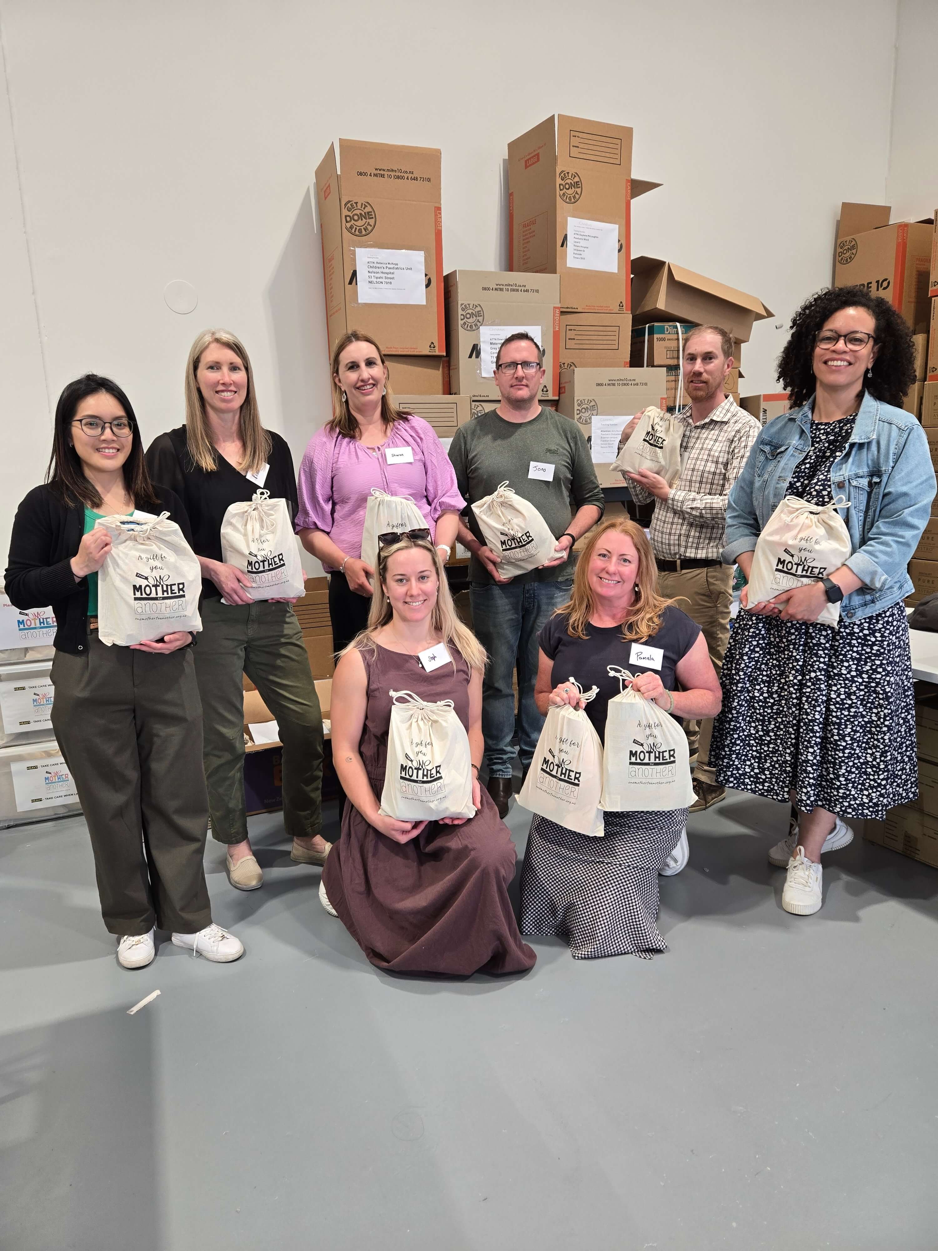 Group of eight people posing with canvas bags labeled 'A gift for you Mother Another' in a warehouse with stacked cardboard boxes.