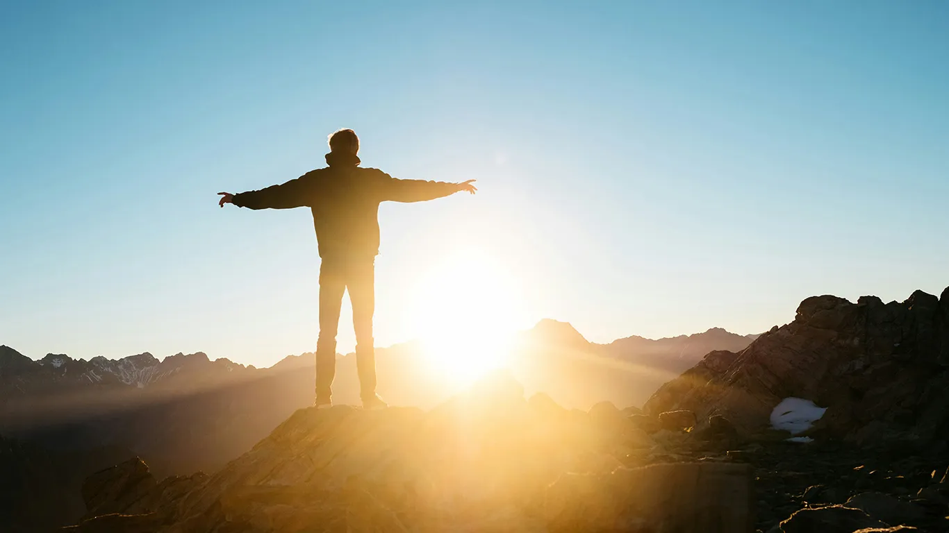 Person steht mit ausgestreckten Armen auf einem felsigen Gipfel bei Sonnenuntergang.