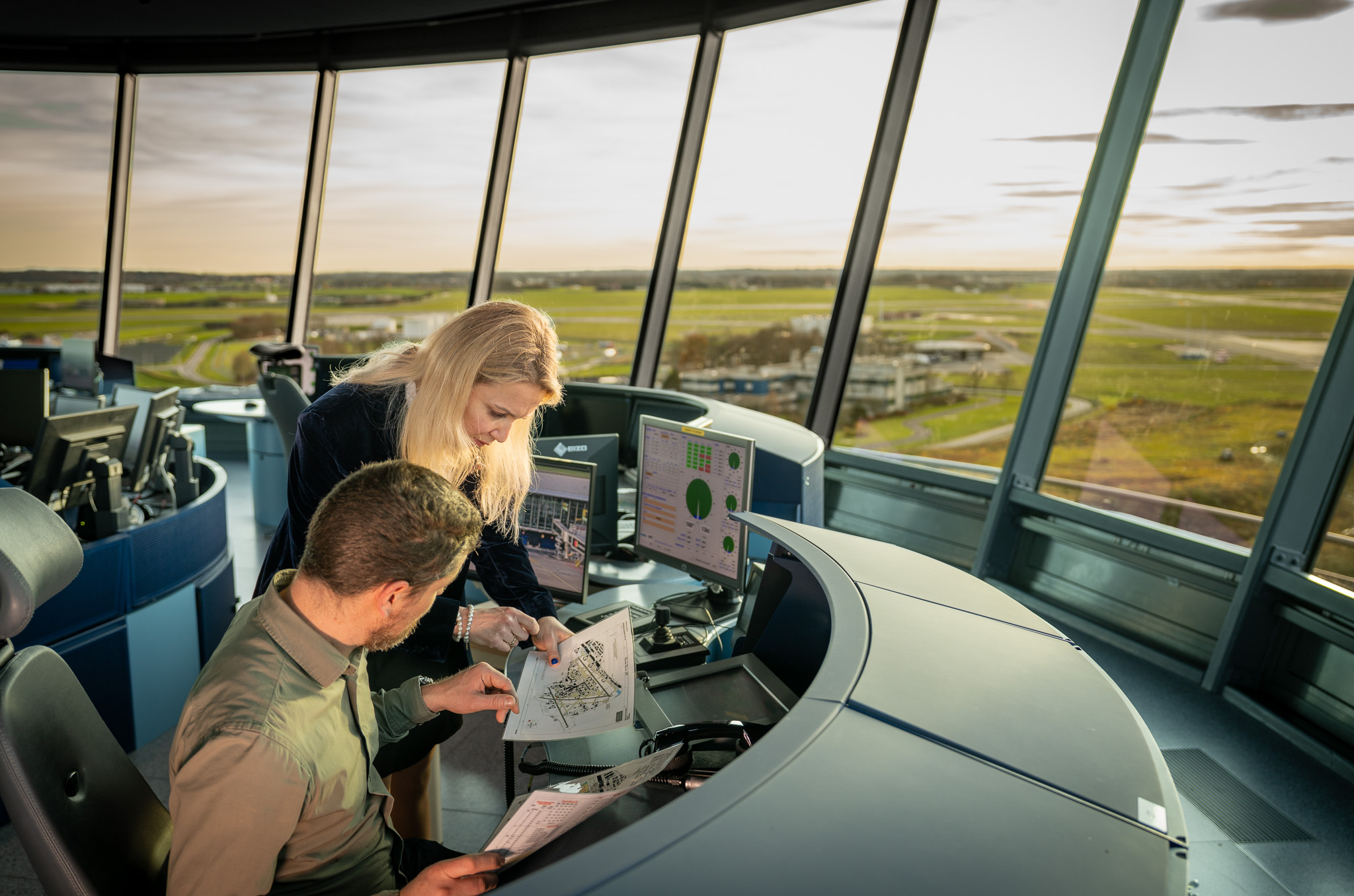 Two air traffic controllers reviewing airport diagrams inside an air traffic control tower with a large window overlooking the airfield.
