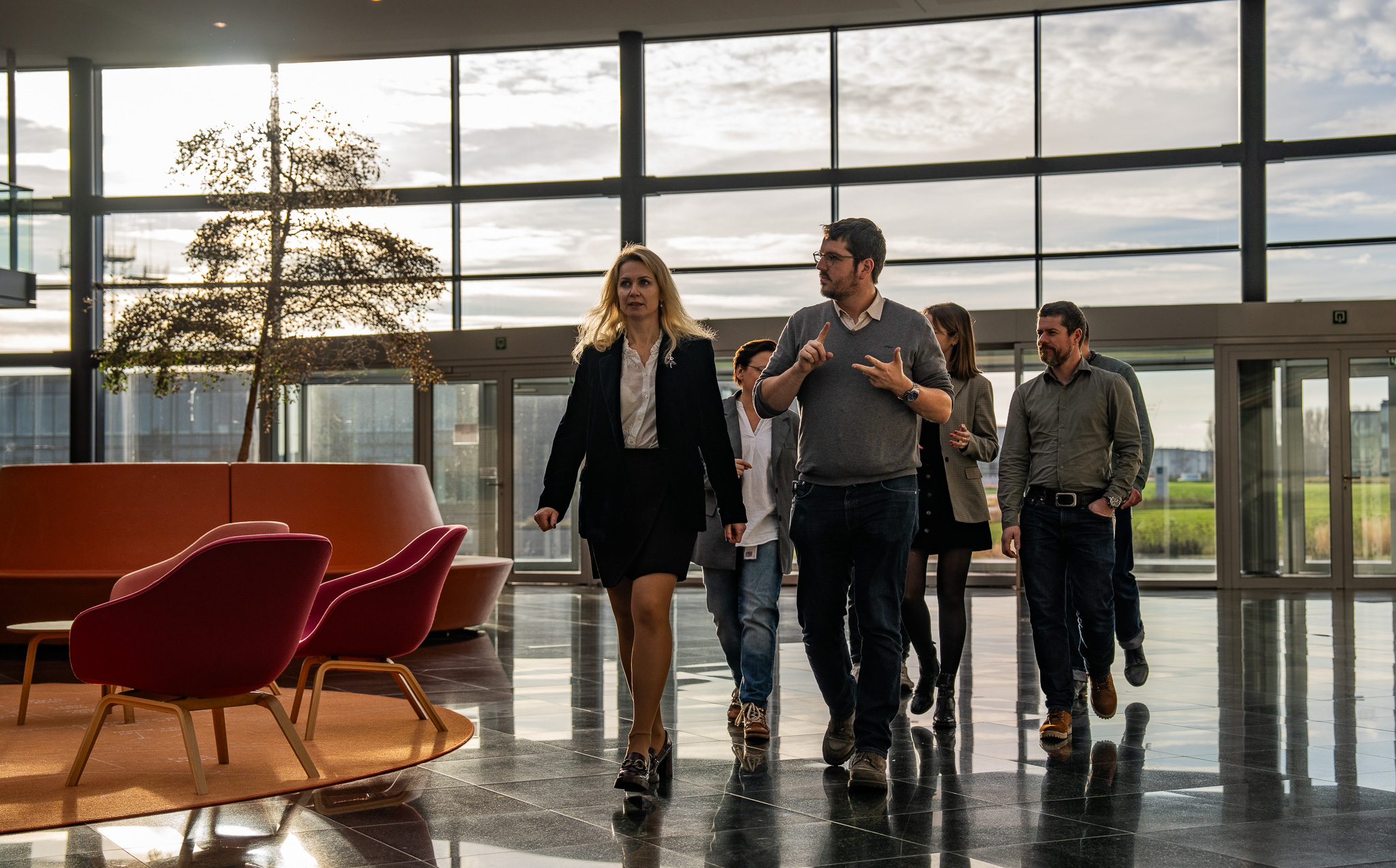 A diverse group of six business professionals walking and talking inside a modern office lobby with large windows and contemporary furniture.