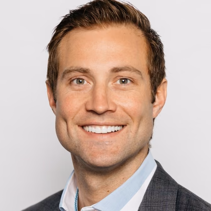 Smiling man with short brown hair wearing a gray suit jacket and light blue shirt.