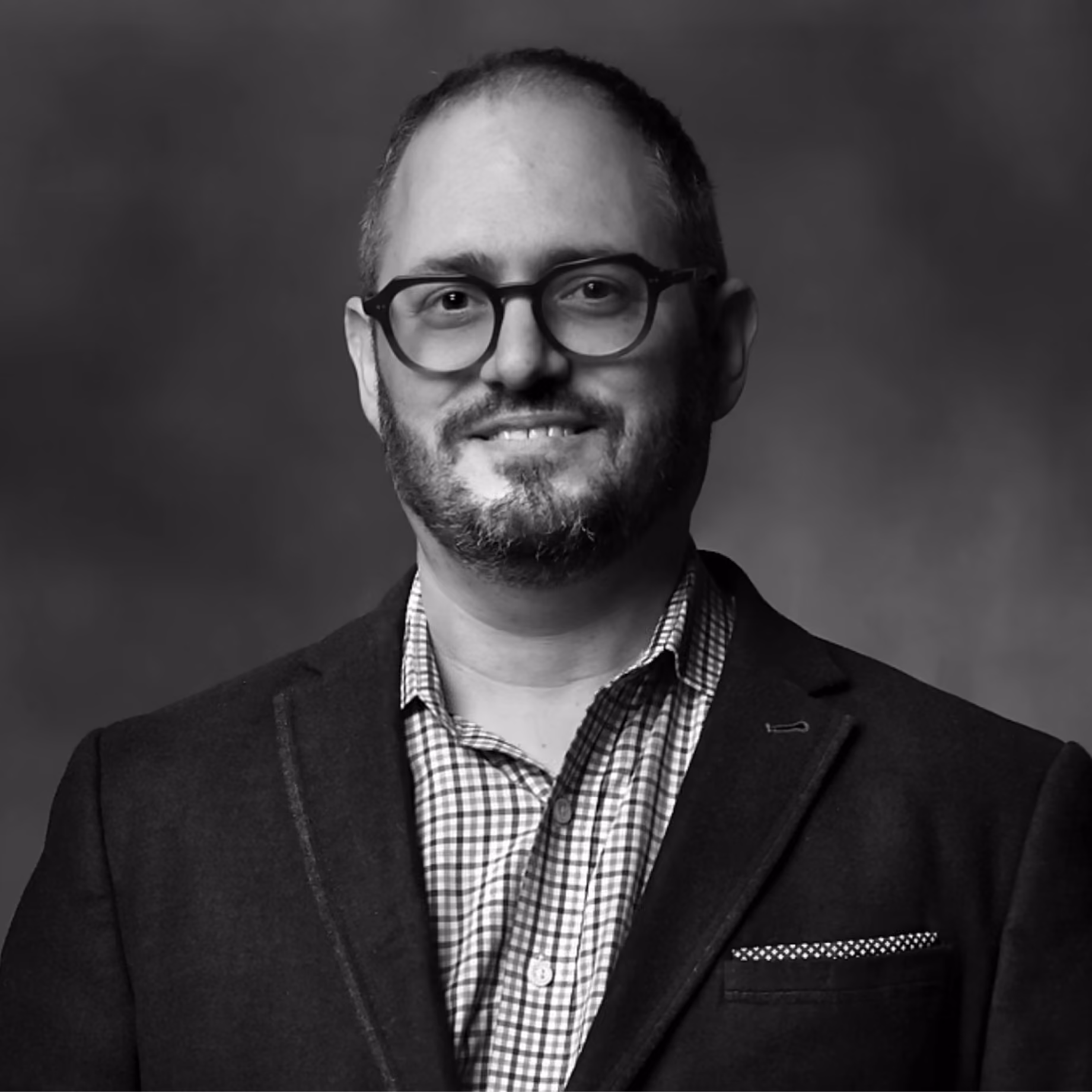 Smiling man with glasses, beard, and checked shirt wearing a dark blazer against a plain background.