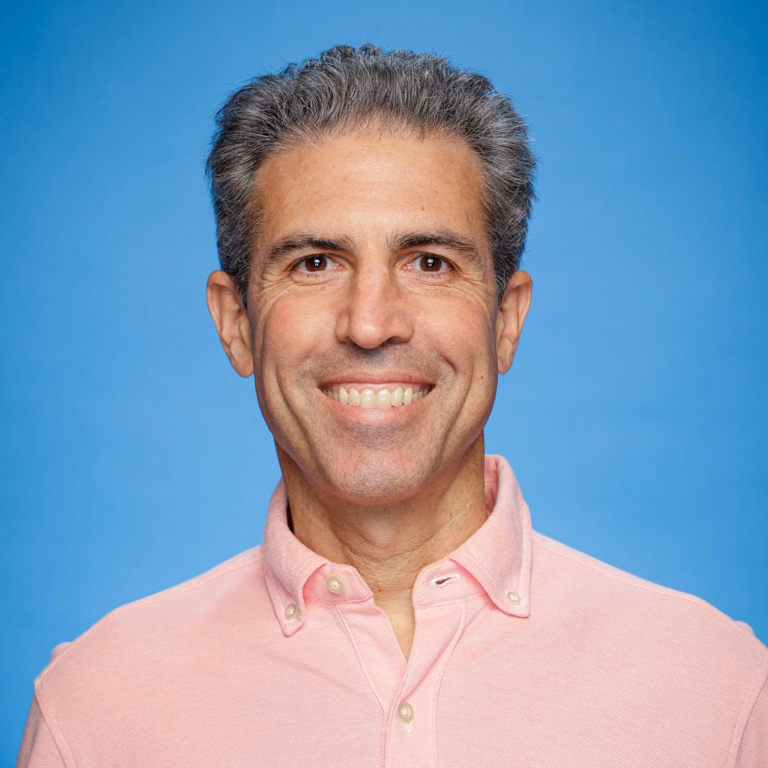 Smiling middle-aged man with gray hair wearing a pink button-up shirt on blue background.