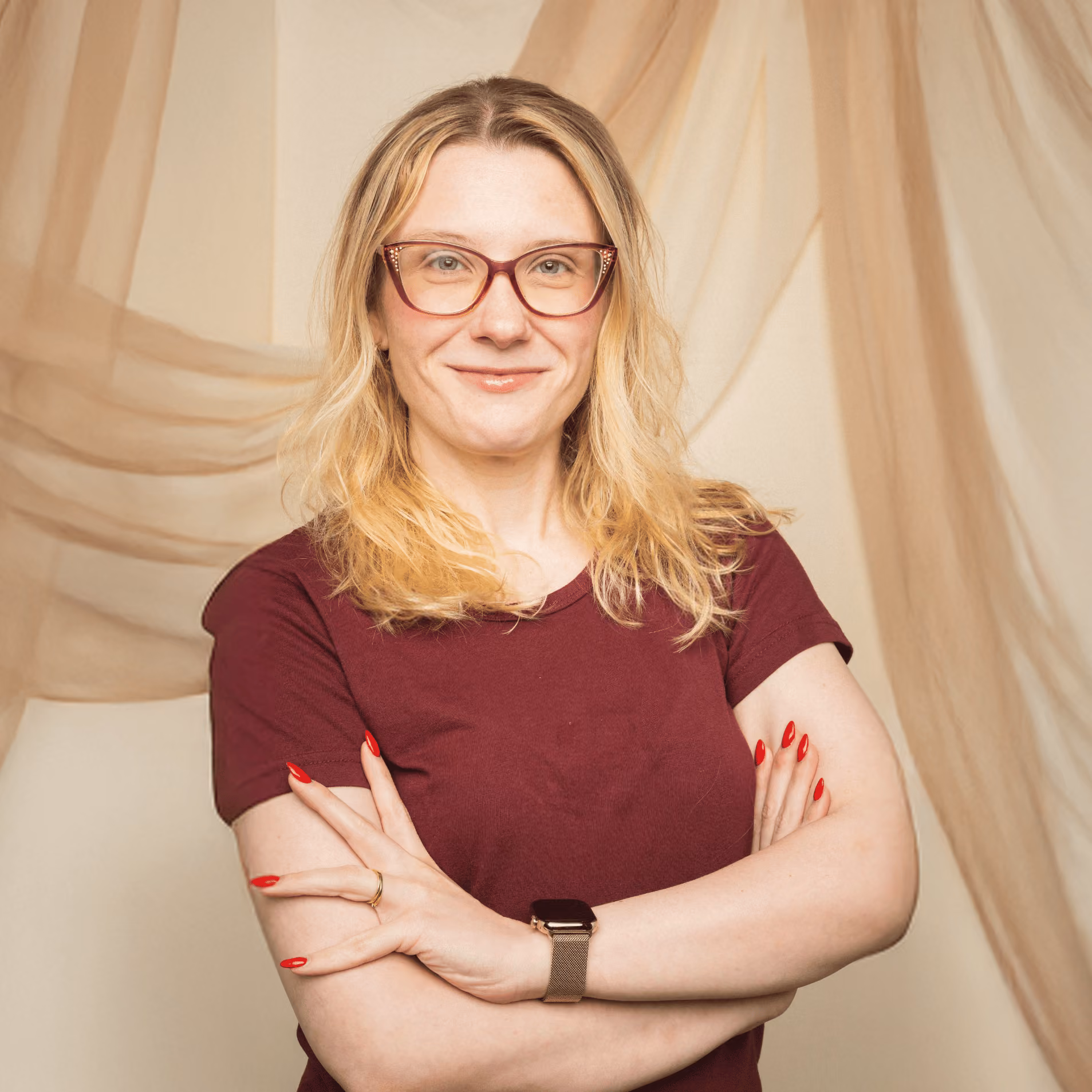 Smiling woman with blond hair, glasses, and red nails wearing a burgundy shirt with arms crossed.