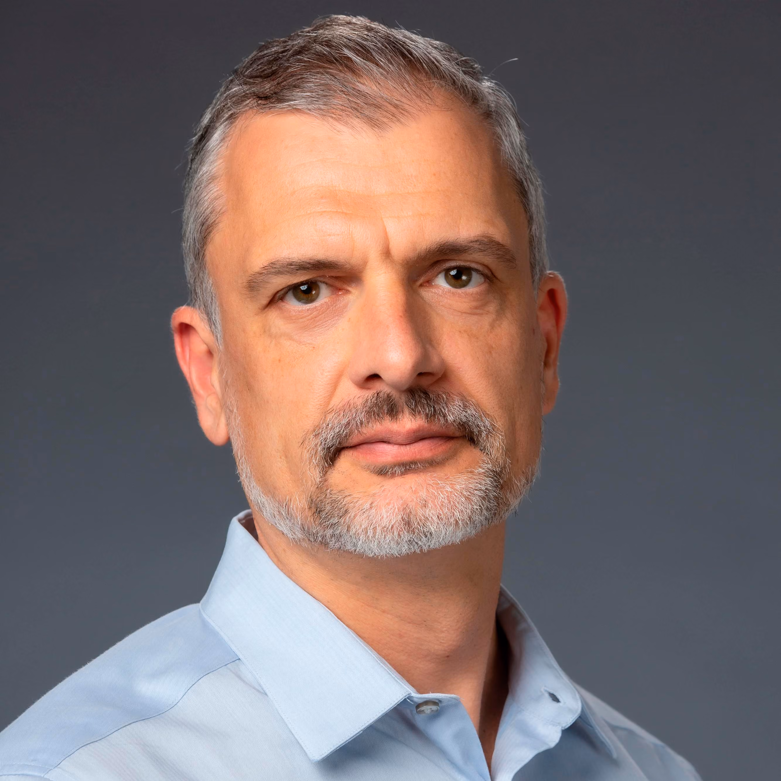 Middle-aged man with gray hair and beard wearing a light blue collared shirt against a gray background.