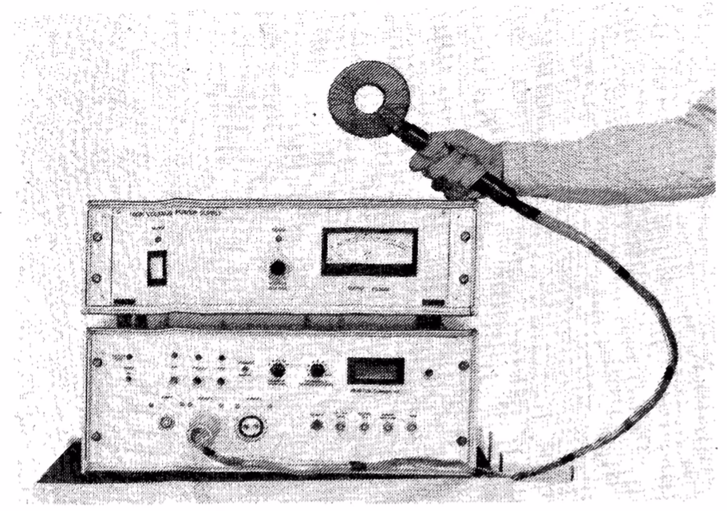 Hand holding a circular probe connected by a cable to vintage electronic testing equipment with dials and meters.