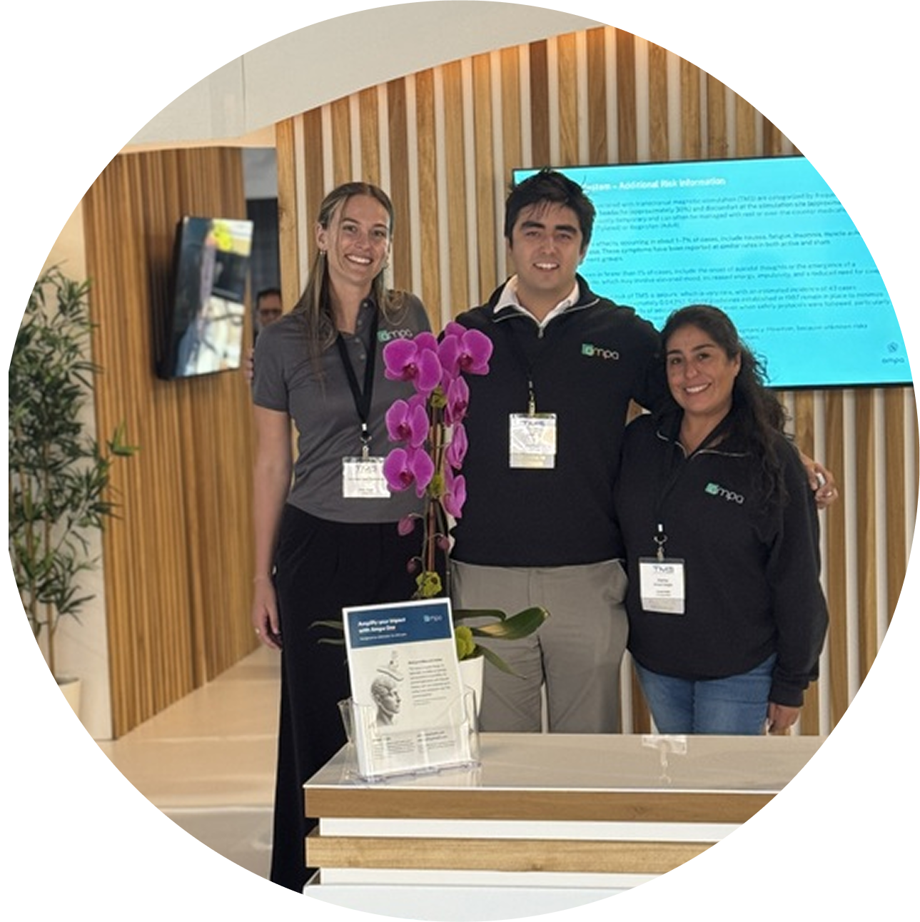 Three people wearing matching shirts stand behind a counter with a purple orchid and brochures.