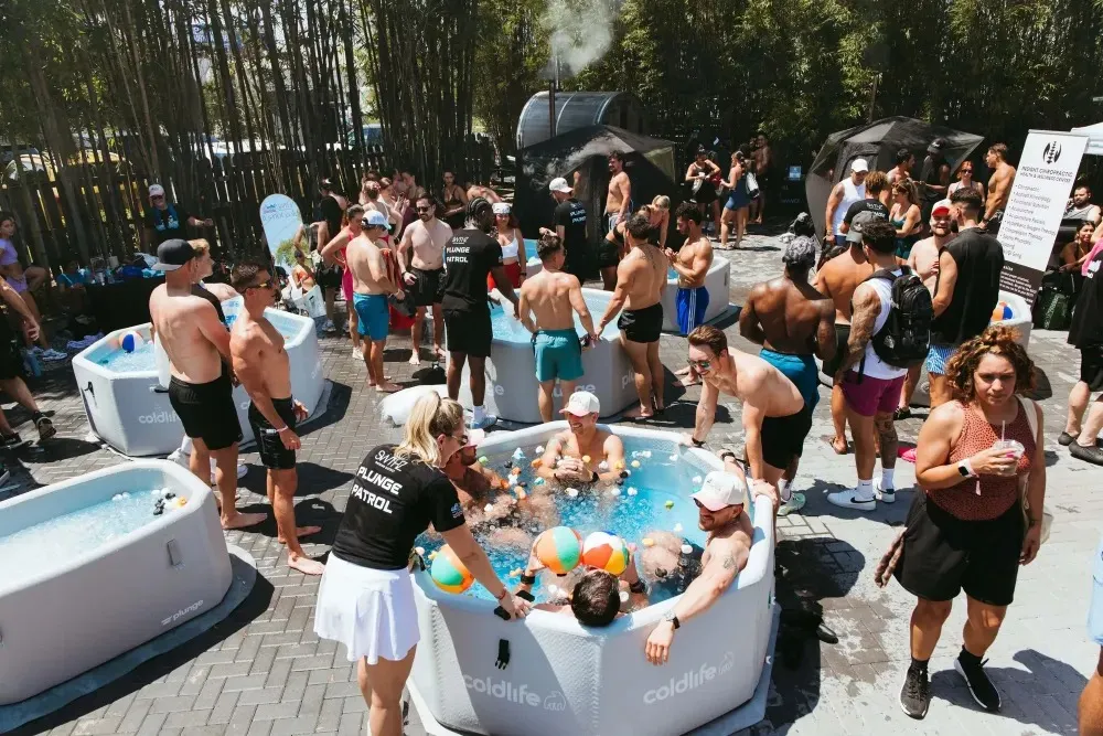 Group of people gathered around and inside multiple portable cold plunge tubs outdoors on a sunny day with trees in the background.