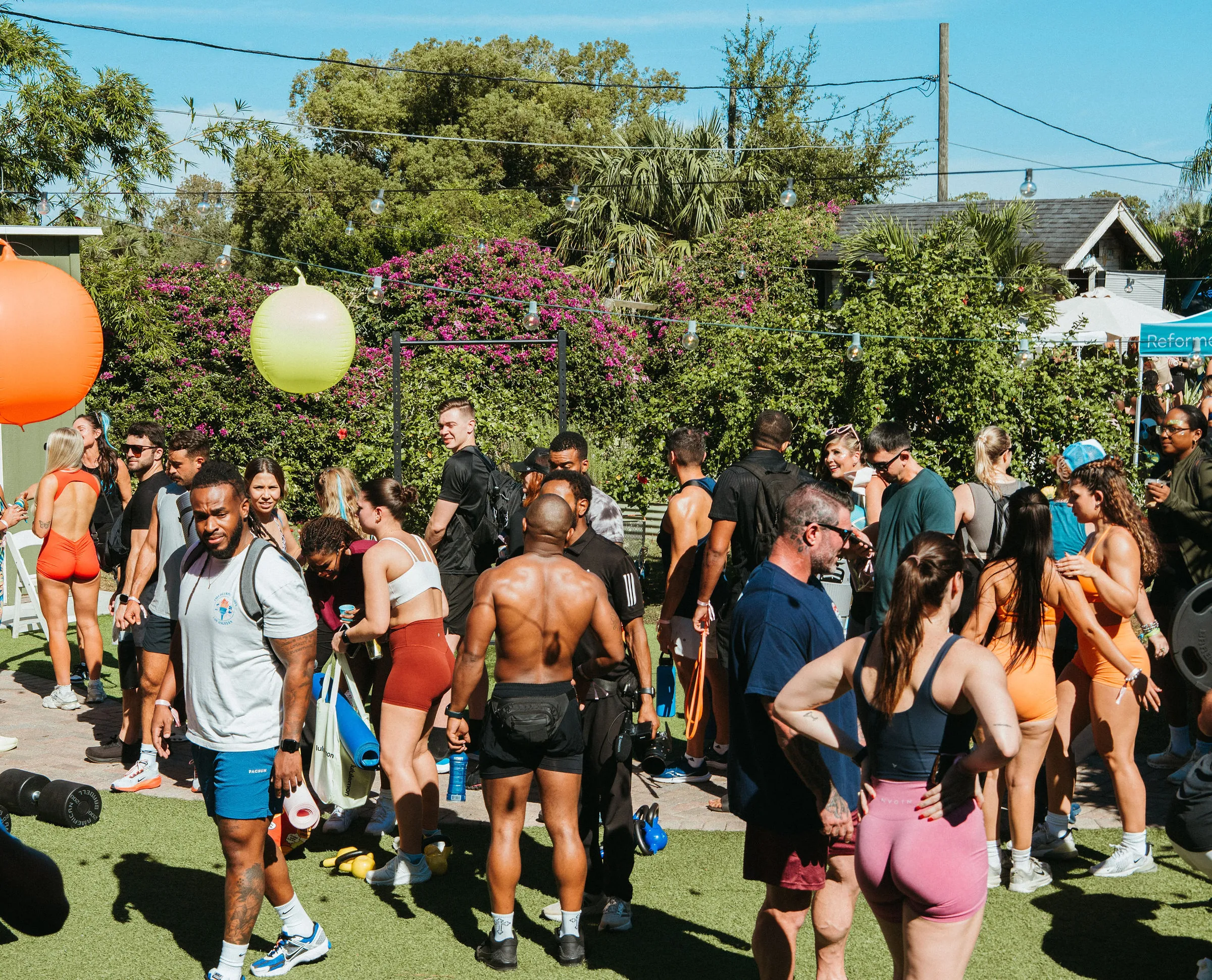 Group of people gathered outdoors on a sunny day, many wearing athletic clothing, with greenery and string lights in the background.