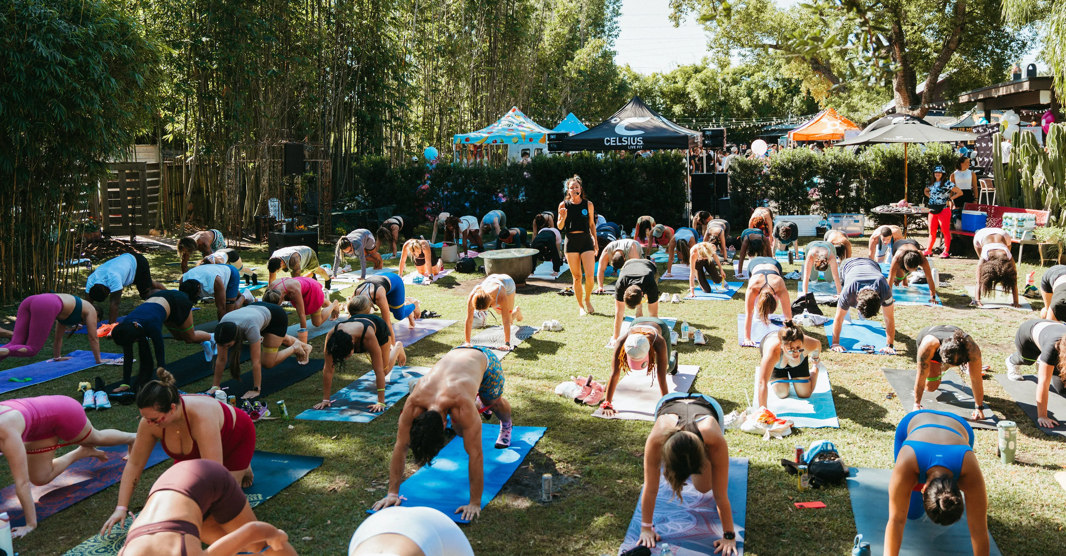 Group of people doing outdoor yoga on mats in a sunny park with a female instructor standing among them.