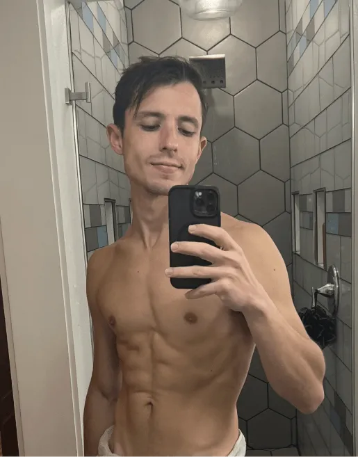 Shirtless young man taking a selfie in a modern bathroom with hexagonal tiles.