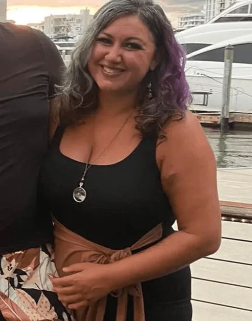 Smiling woman with curly hair dyed grey and purple, wearing a black tank top and brown skirt with a bow, standing near a dock with boats in the background.