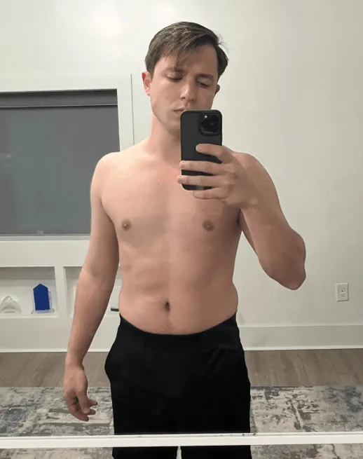 Shirtless young man with short brown hair taking a mirror selfie holding a smartphone in a room with a window and patterned rug.