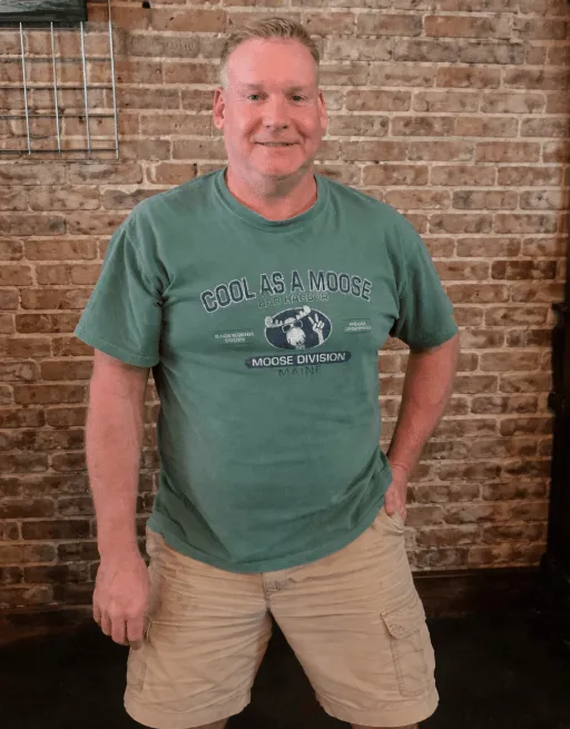 Smiling man wearing a green 'Cool As A Moose Maine' t-shirt and beige shorts standing in front of a brick wall.