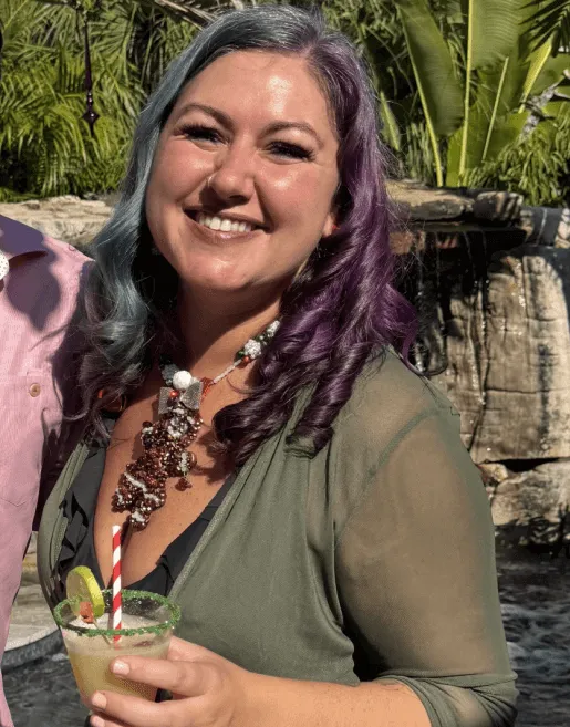 Smiling woman with blue and purple curly hair wearing a green outfit and holding a cocktail with a lime and striped straw outdoors.