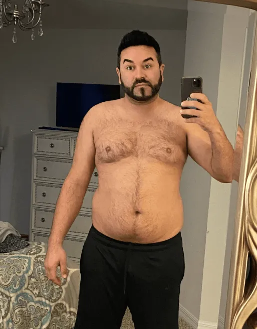 A man with a trimmed beard and chest hair taking a shirtless mirror selfie in a bedroom.