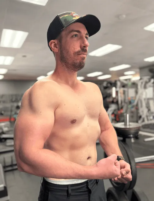 Shirtless man wearing a camouflage baseball cap flexing his arm muscles in a gym.